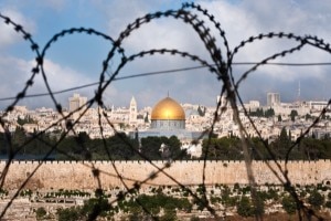 A view of the Old City of Jerusalem, including the Dome of the Rock through coils of razor wire, illustrating the Holy Land’s history of division and conflict. © Ryan Rodrick Beiler | Shutterstock